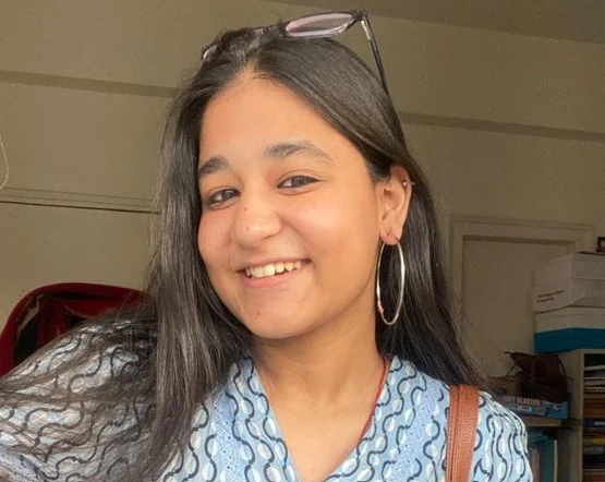 Smiling woman with glasses on head and hoop earrings in a patterned blue top. Background shows shelves and boxes, conveying a casual setting.