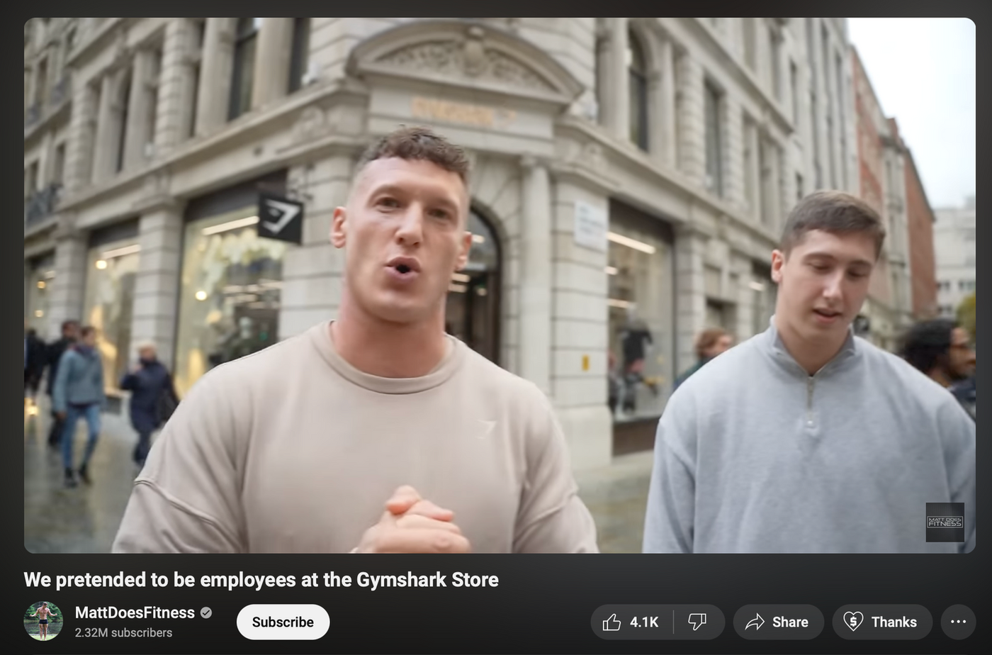 Two men stand outside a Gymshark store on a city street. One speaks animatedly, while people walk by. Store sign and logo visible.