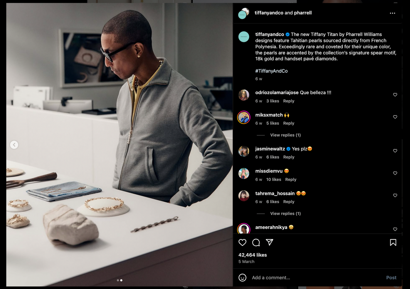 Pharrell Williams examines jewelry on display in a modern, stylish store. Background features framed pictures and neutral tones.
