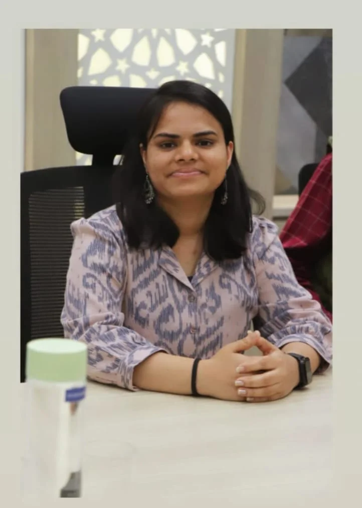Woman in patterned blouse sits at a table, hands folded. Office setting with geometric background; calm expression. Water bottle visible.