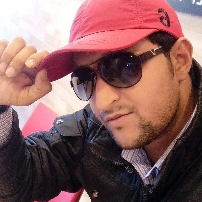 Man in a red cap and sunglasses adjusts the brim, wearing a dark jacket indoors. Background is softly lit, mood is casual.