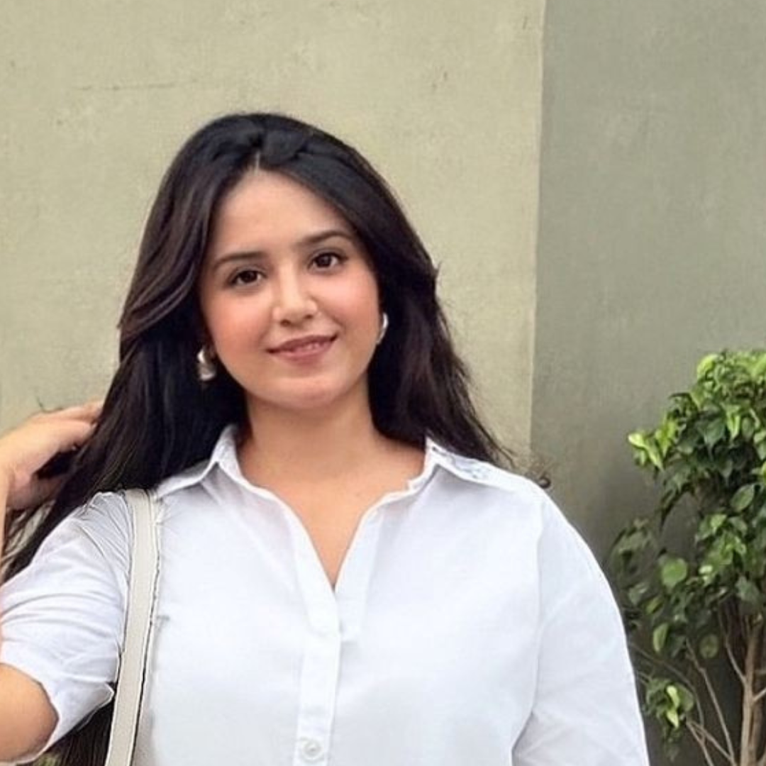 Smiling woman in a white shirt stands outdoors, holding a bag. Background features a light wall and green plant. Casual and cheerful vibe.