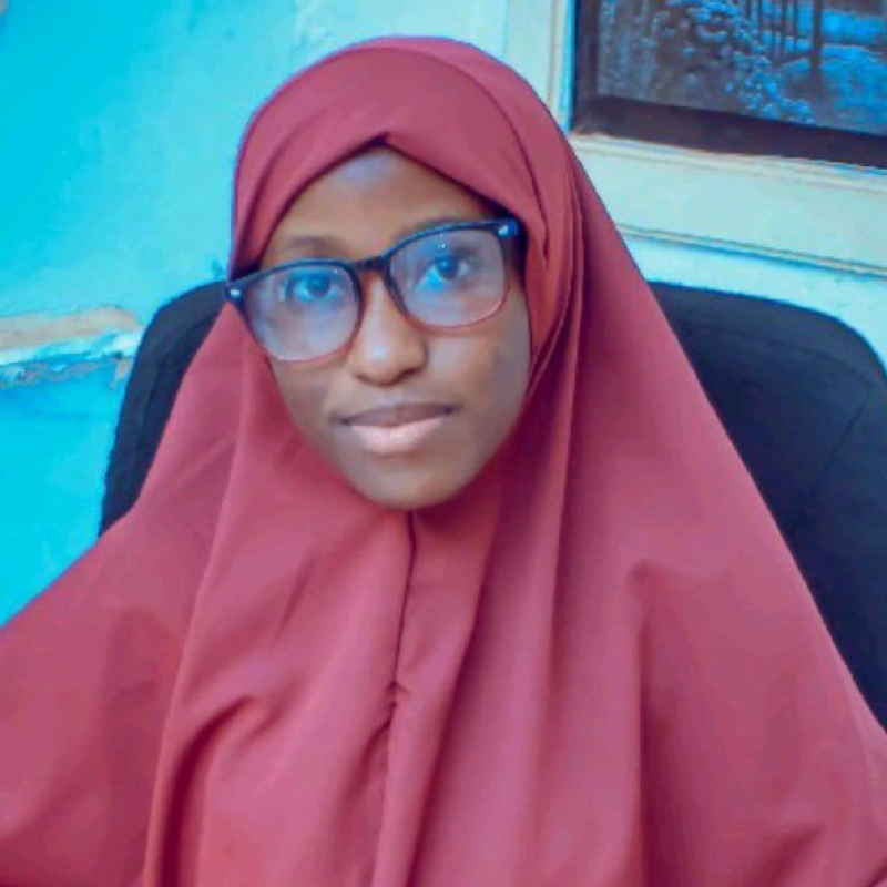 Person in a maroon hijab and glasses, seated indoors against a blue background, with a calm expression.